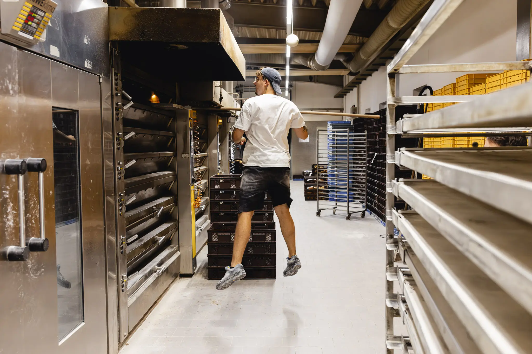 Bäcker springt hoch, um in den geöffneten Backofen in der Backstube zu schauen. Um ihn herum stehen Backblechwagen und Kisten für Backwaren.