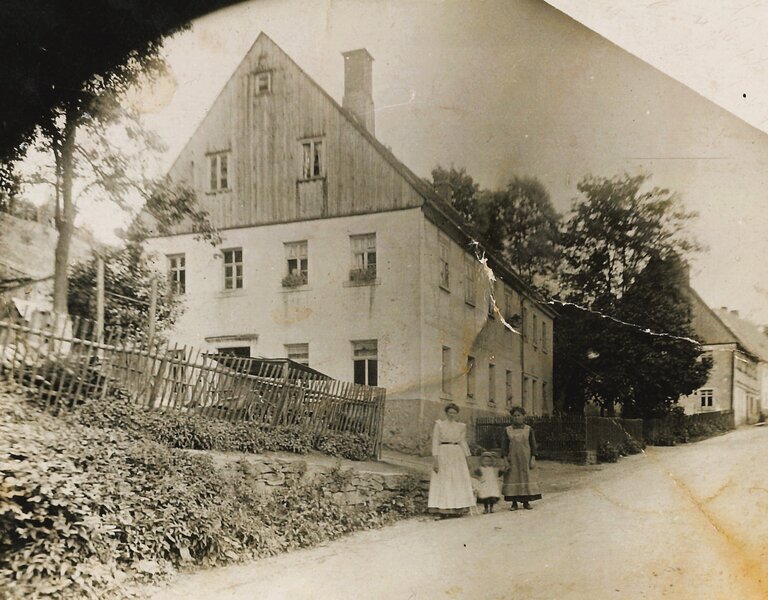Historische Schwarz-Weiß-Aufnahme eines Hauses, vor dem zwei Frauen mit einem kleinen Mädchen an den Händen stehen.