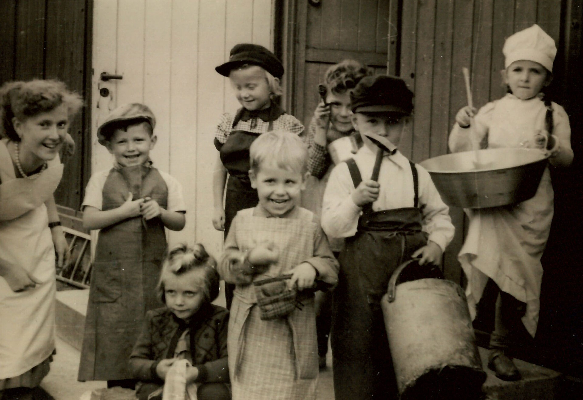 Historische Schwarz-Weiß-Aufnahme von lachenden Kindern in Schürzen und Bäckermützen mit Töpfen und Küchenutensilien.